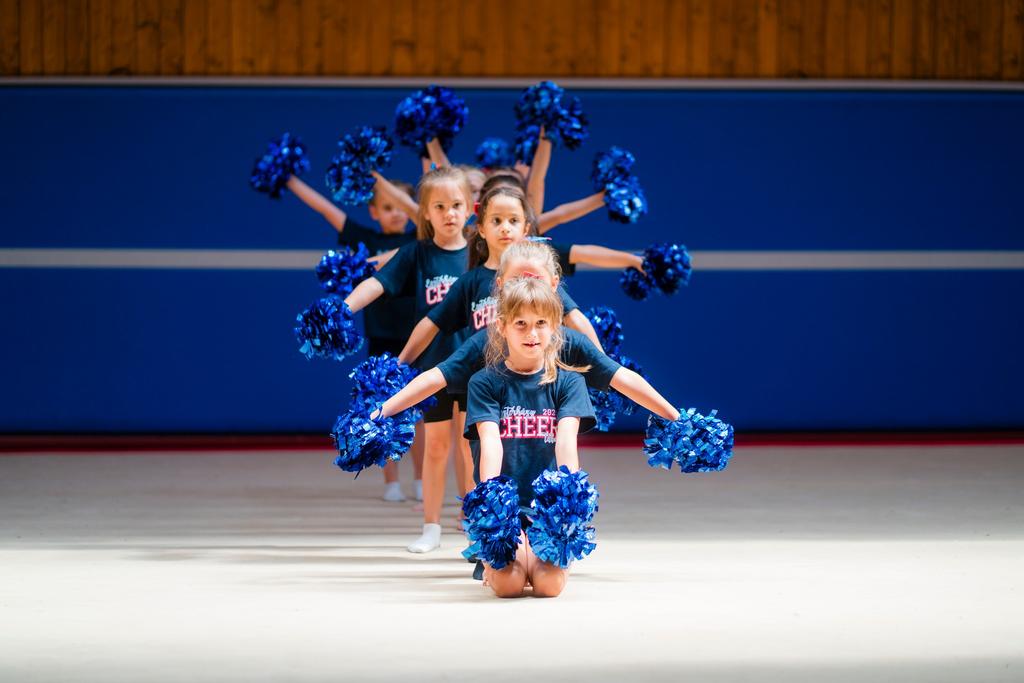 Egy sor cheerleader lány tintákkal, kék pom-ponokat tartva a kezükben egy sportcsarnokban.